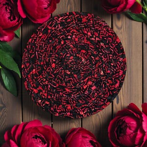 Könnte beinhalten: Ein runder, schwarz-roter gewebter Platzdeckchen mit Spiralmuster. Der Platzdeckchen ist von leuchtend roten Pfingstrosen und grünen Blättern umgeben, vor einem Holzgrund.