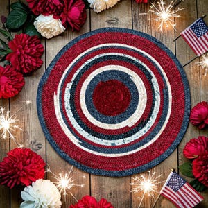 Könnte beinhalten: Ein runder geflochtener Teppich mit konzentrischen Kreisen in Rot, Weiß und Blau. Der Teppich ist von roten und weißen Blumen, amerikanischen Flaggen und Wunderkerzen umgeben, was ein patriotisches Thema schafft. Der Teppich liegt auf einer Holzoberfläche.