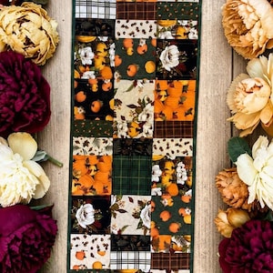 May include: A rectangular patchwork table runner featuring a grid of fabric squares with patterns of orange pumpkins, white flowers, green leaves, and dark plaid designs, bordered by a dark green trim and flanked by clusters of burgundy and cream peonies.