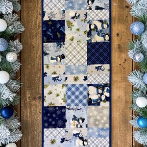 May include: A rectangular patchwork table runner featuring a variety of blue, white, and grey fabric squares with patterns of snowmen, snowflakes, plaid, and holly, flanked by two frosted evergreen garlands decorated with blue and white ornaments.