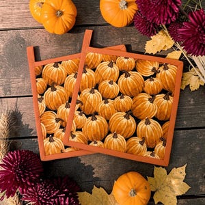 May include: Two square fabric pot holders featuring a repeating pattern of orange pumpkins on a black background with solid orange fabric trim and a small hanging loop on the top edge of each piece, designed for kitchen use and heat protection.
