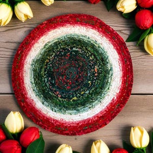 Op de afbeelding: Een ronde, handgemaakte vloerkleed met concentrische cirkels van rode, witte en groene stof. Het vloerkleed is omgeven door gele tulpen en rode bessen op een houten oppervlak. Het midden van het vloerkleed heeft een gedetailleerd patroon.