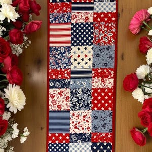 May include: A rectangular patchwork table runner with a red border. The runner features a variety of patriotic patterns in red, white, and blue, including stars, stripes, and floral designs. Red roses and white flowers frame the runner.