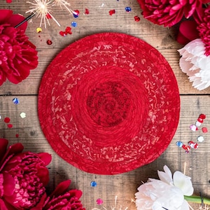 Peut inclure: Un set de table circulaire rouge vif avec un motif texturé est posé sur une surface en bois rustique. L'image est ornée de pivoines rouges et blanches, de confettis et de feux d'artifice, créant une ambiance festive.