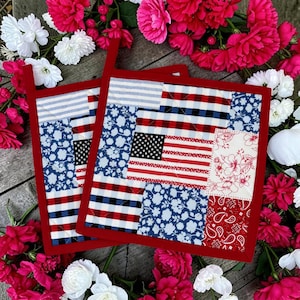 Puede incluir: Dos salvamanteles cuadrados acolchados con borde rojo. El diseño de patchwork presenta la bandera estadounidense, estampados florales y un estampado de cachemira rojo. Los salvamanteles están rodeados de flores rosas y blancas.