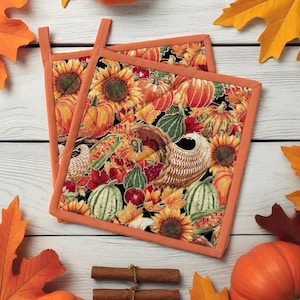 Può includere: Due presine quadrate con un tema del raccolto autunnale. Il tessuto presenta girasoli, zucche, zucche e una cornucopia su sfondo nero. Le presine hanno bordi arancioni e anelli per appenderle. Bastoncini di cannella e foglie autunnali sono anche nell'immagine.