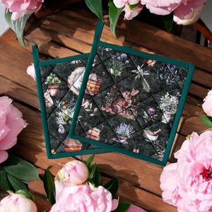 Könnte beinhalten: Zwei grüne, gesteppte Topflappen mit einem floralen und tierischen Muster. Der Druck zeigt ein Kaninchen, einen Vogel, einen Bienenstock und verschiedene Blumen. Die Topflappen haben einen dunkelgrünen Rand.