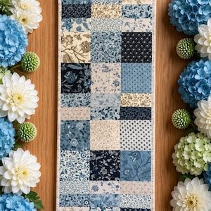 May include: A rectangular table runner featuring a patchwork design of various fabric squares in shades of navy blue, light blue, and cream. The fabric squares display diverse floral, botanical, and geometric patterns arranged in a grid layout.