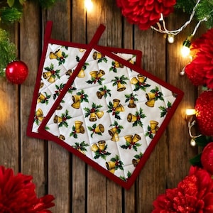 Könnte beinhalten: Zwei weiße Topflappen mit rotem Rand und einem Weihnachtsglocken- und Stechpalmenmuster. Die Topflappen liegen auf einer Holzoberfläche mit roten Blumen und Weihnachtslichtern.