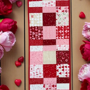 May include: A patchwork table runner with a red border, featuring squares of various red and pink patterns, including hearts and floral designs. The runner is surrounded by pink and red peonies and heart-shaped candies.