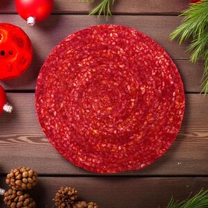Könnte beinhalten: Ein runder, roter Tischset mit Spiralmuster, umgeben von Weihnachtsschmuck und Tannenzapfen auf einer Holzoberfläche. Das Tischset hat eine leuchtend rote Farbe und eine strukturierte Optik. Das Bild erzeugt eine festliche Urlaubsstimmung.