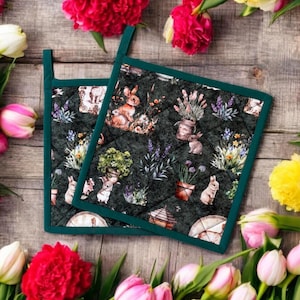 Könnte beinhalten: Zwei grüne, gesteppte Topflappen mit einem Blumen- und Hasendesign. Die Topflappen haben einen dunkelgrünen Hintergrund mit einem hellgrünen Steppmuster. Das Design zeigt Hasen, Blumen und Pflanzen.