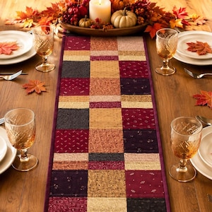 May include: A rectangular table runner featuring a patchwork design of alternating squares in shades of burgundy, dark purple, and gold. The fabric squares display subtle patterns and textures, bordered by a solid dark purple trim along the edges.