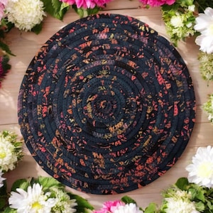 Pode incluir: Um descanso de copo redondo, preto e branco, tecido com um padrão floral rosa e dourado. O descanso de copo é feito de corda e tem um design circular.