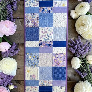 May include: A rectangular quilted table runner featuring a patchwork design of various blue, purple, and white floral and geometric fabric squares. The runner is bordered with a solid purple fabric trim and displays a repeating grid pattern.
