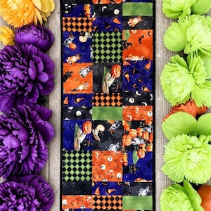 May include: A rectangular fabric table runner featuring a patchwork design of Halloween-themed patterns in orange, purple, black, and green, flanked by vertical rows of large, artificial purple and lime green peony-style flowers.