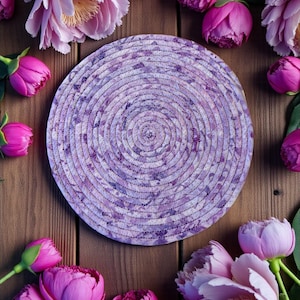 Pode incluir: Uma base redonda, roxa e branca, com um design em espiral. A base é cercada por peônias rosa e roxas em uma superfície de madeira. A base é feita de um material tecido.
