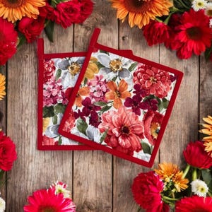 May include: Two red and white floral print pot holders with a red trim. The pot holders are on a wooden surface surrounded by a border of red, orange, and yellow flowers.