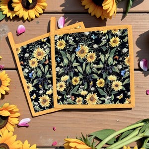 May include: Two square pot holders with a black background, adorned with a sunflower and butterfly pattern, and trimmed with yellow fabric. The pot holders are placed on a wooden surface, surrounded by sunflowers and petals.