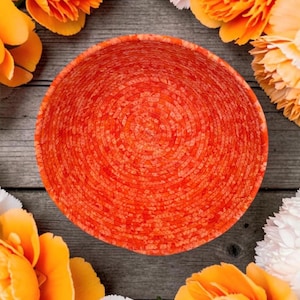 May include: A circular bowl constructed from coiled rope in a vibrant orange hue. The rope features a mottled texture with varying shades of red and orange, creating a consistent spiral pattern that radiates from the center to the outer rim.