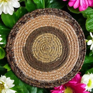 May include: A round, brown, beige, and white woven coaster with a concentric circle pattern. The coaster is surrounded by green leaves and pink flowers.