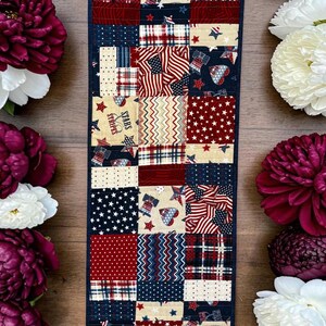 Pode incluir: Um caminho de mesa patriótico com um design de patchwork em vermelho, branco e azul. O caminho de mesa inclui estrelas, listras e motivos de bandeiras. O caminho de mesa é cercado por flores bordô e brancas. Mede cerca de 100 cm.