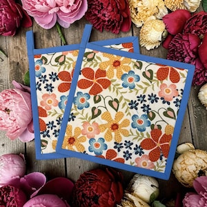 May include: Two square quilted pot holders featuring a cream-colored fabric with a repeating floral pattern in shades of orange, yellow, light blue, and dark blue. Each pot holder is finished with a solid blue fabric binding and a small hanging loop.