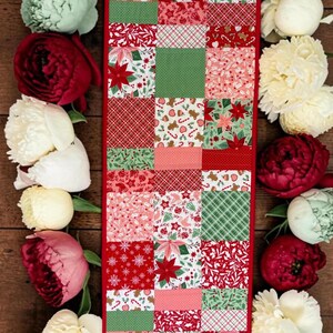 Può includere: Un runner da tavola rettangolare caratterizzato da un design patchwork con vari tessuti a tema natalizio nei colori rosso, verde e bianco. I motivi includono stelle di Natale, omini di pan di zenzero, bastoncini di zucchero e fiocchi.