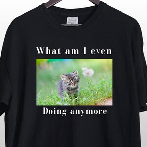 May include: Black t-shirt with white text that reads "What am I even doing anymore."  A photo of a small kitten looking at a dandelion is centered on the shirt.