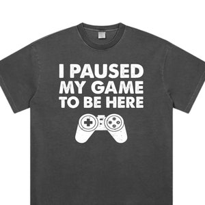 Puede incluir: Camiseta unisex gris jaspeado oscuro con el texto "I PAUSED MY GAME TO BE HERE" y un gráfico de mando blanco. Un estilo casual y cómodo.
