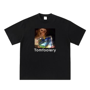 Puede incluir: Camiseta negra con una imagen de un mono bebiendo de una caja de jugo. El texto "Tomfoolery" está impreso debajo de la imagen.