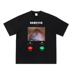 May include: Black t-shirt featuring a graphic of a hamster making a phone call on a screen. The text "HAMSTER is calling" is above the image. The phone screen has buttons that say "Remind Me", "Message", "Decline", and "Accept".