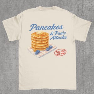 May include: A cream-coloured t-shirt with a design featuring a stack of pancakes with butter and syrup, blueberries, and the text "Pancakes & Panic Attacks" in blue script. A red oval reads "buy one get one".