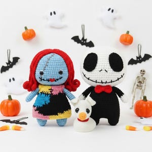 May include: Three crocheted dolls featuring a blue-skinned character with red yarn hair, a white-headed character in a black suit with a red bow tie, and a small white dog with a red collar, all displayed with miniature Halloween decorations.