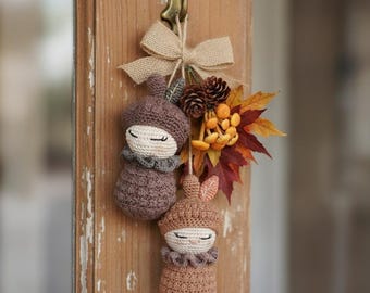 May include: Two crocheted acorn-shaped dolls with closed eyes, one dark brown and one light brown, hanging from a metal hook. The dolls are tied with a burlap bow and decorated with two small pinecones, orange berries, and autumn-colored leaves.