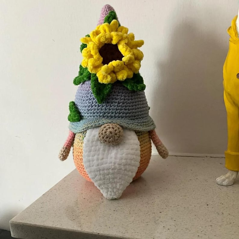 May include: A handcrafted crochet gnome featuring a light blue pointed hat adorned with a large, textured yellow sunflower and green leaves. The gnome has a round beige nose, a long white fuzzy beard, and a body with a gradient orange-to-pink finish.