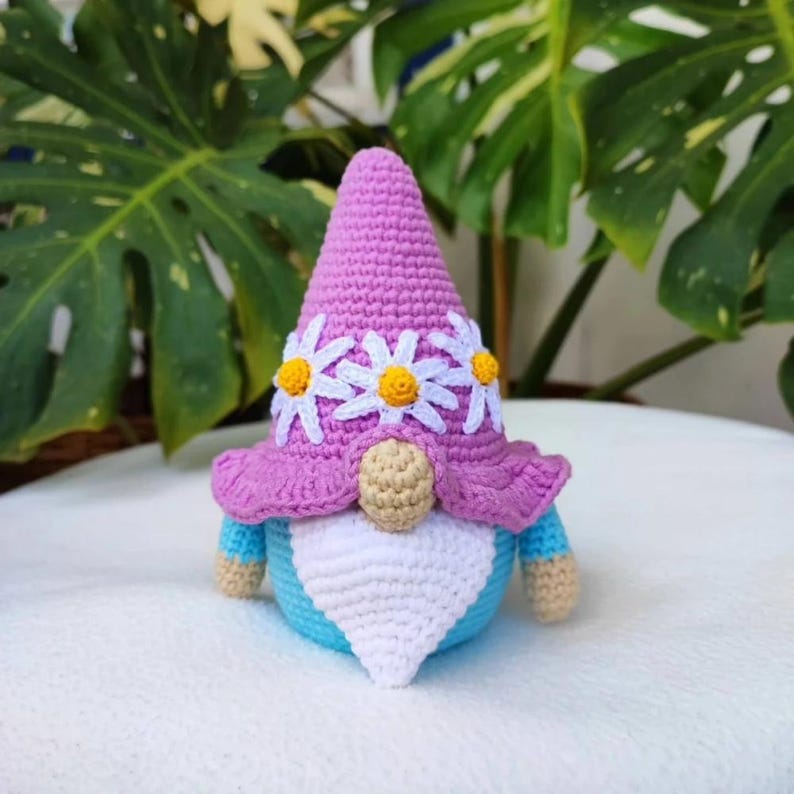 May include: A crochet gnome figurine featuring a tall, pointed purple hat decorated with three white daisy flowers with yellow centers. The gnome has a beige nose, a white triangular beard, and a light blue body with beige-colored hands.