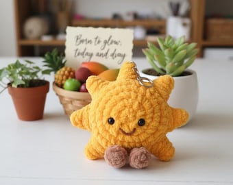 May include: A yellow crocheted sun keychain with a smiling face, black bead eyes, and small brown feet. The plush sun features a metal keyring attached to the top and is crafted with a soft, textured yarn finish, showcasing a cheerful design.