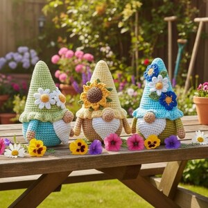 May include: Three crocheted gnome figurines sit on a wooden table. The left gnome is light green and blue with white daisies. The center gnome is yellow and tan with a large sunflower. The right gnome is blue and green with blue and white flowers.