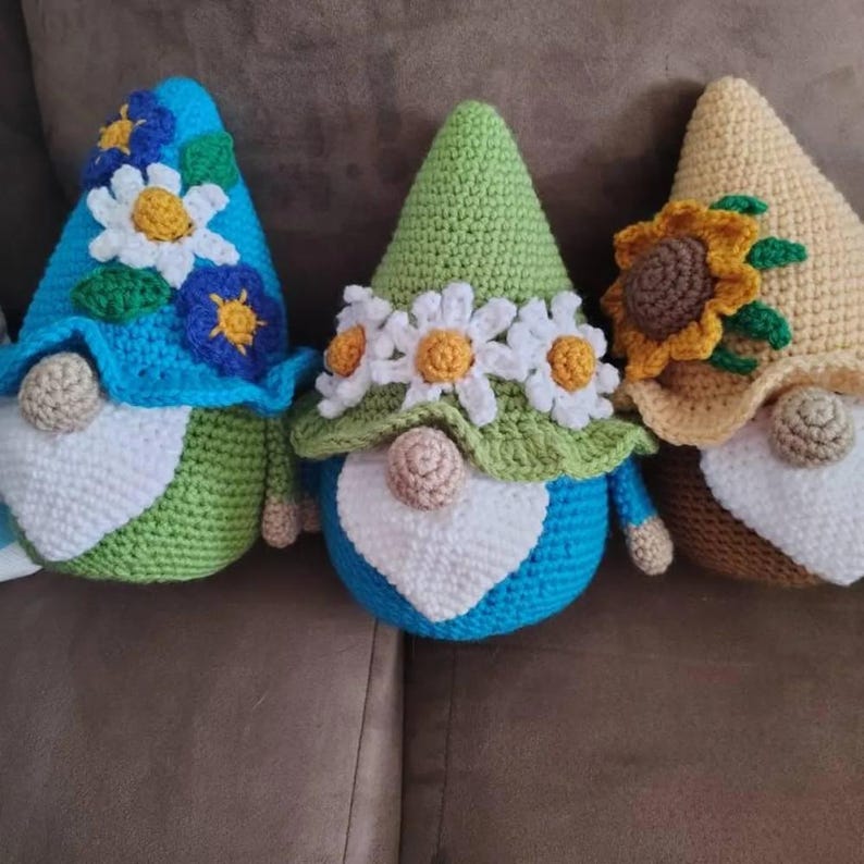 May include: Three handmade crochet gnomes featuring pointed hats adorned with floral appliques. The left gnome has a blue hat with white and blue flowers, the center has a green hat with white daisies, and the right has a tan hat with a sunflower.