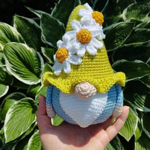 May include: A handmade crochet gnome featuring a bright yellow pointed hat adorned with three white and yellow daisy flowers. The gnome has a light blue body, a white beard, and a small round beige nose, crafted with detailed yarn stitching.