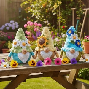 May include: Three crocheted gnomes with pointed hats, each featuring a white beard and a round nose. The left gnome has a green hat with white daisies, the center has a yellow hat with a sunflower, and the right has a blue hat with blue and white flowers.