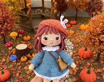 May include: A crochet doll with reddish-brown hair, wearing an orange beret with a small white bunny accent, a blue long-sleeved dress with a white collar, a small tan crossbody bag, white socks, and brown shoes with tan details.