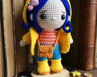 May include: A crocheted doll with blue hair, a yellow hooded jacket, a striped orange and white shirt, blue pants, and yellow boots. The doll wears a small orange crossbody bag and a pink flower accessory in its hair, standing on a wooden base.
