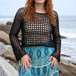 May include: A black, open-weave crochet top with long sleeves. The top is paired with a blue and turquoise patterned skirt with a white trim. The background shows a rocky shoreline and the sea.