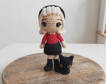May include: A crochet doll with white hair, a red long-sleeved top, a black skirt, and black boots, standing next to a small black crochet cat with yellow eyes. The doll has a round head, dark eyes, and a light skin tone, crafted in a yarn style.