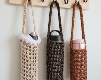 May include: Three crochet water bottle holders in beige, brown, and pink hang from a wooden rack. Each holder has a long strap and a unique design. The beige holder has a fluffy white trim, while the others are solid colors.