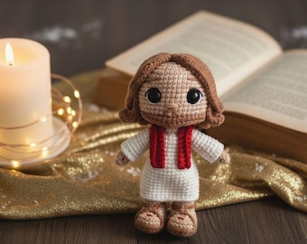 May include: A small, handmade crochet doll featuring a figure with light brown yarn hair and a beard, wearing a white tunic with a red sash draped over the shoulders. The doll has large black safety eyes and is crafted with detailed stitching.