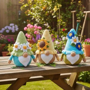 May include: Three crocheted gnome figurines featuring pointed hats adorned with fabric flowers. The left gnome has a light green hat, the center has a yellow hat with a sunflower, and the right has a blue hat with blue and white flowers.