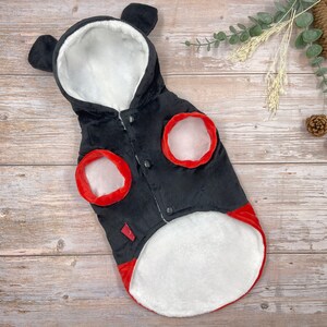 May include: Black dog coat with a hood and bear ears. The coat has red trim around the armholes and bottom, with a white fluffy interior. The coat fastens with black buttons.